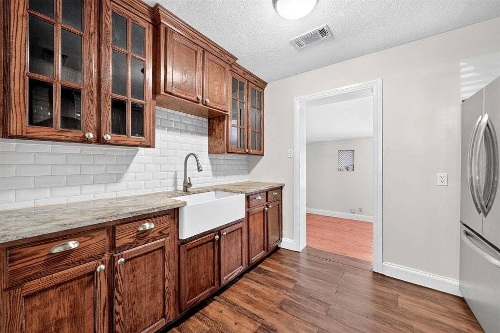 Rich-toned wood cabinetry with glass-front uppers, white subway tile backsplash, white farmhouse sink, granite countertop, and wood-finish flooring