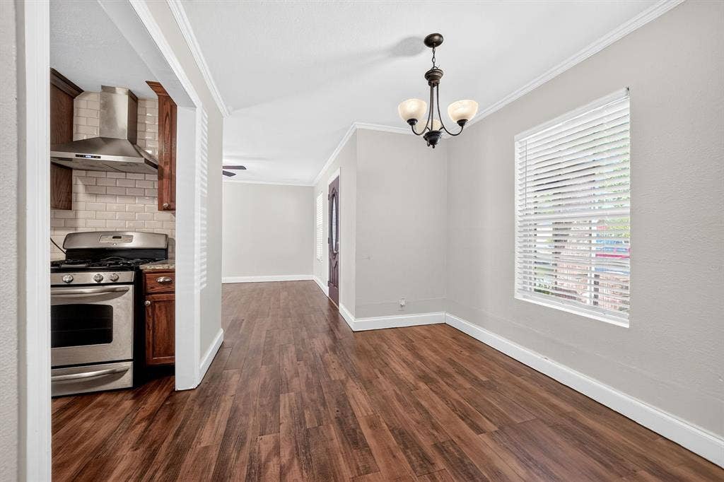 Dining area featuring wood-finish flooring, crown molding, and a three-light chandelier, with a partial view of a kitchen equipped with a stainless steel range and subway tile backsplash