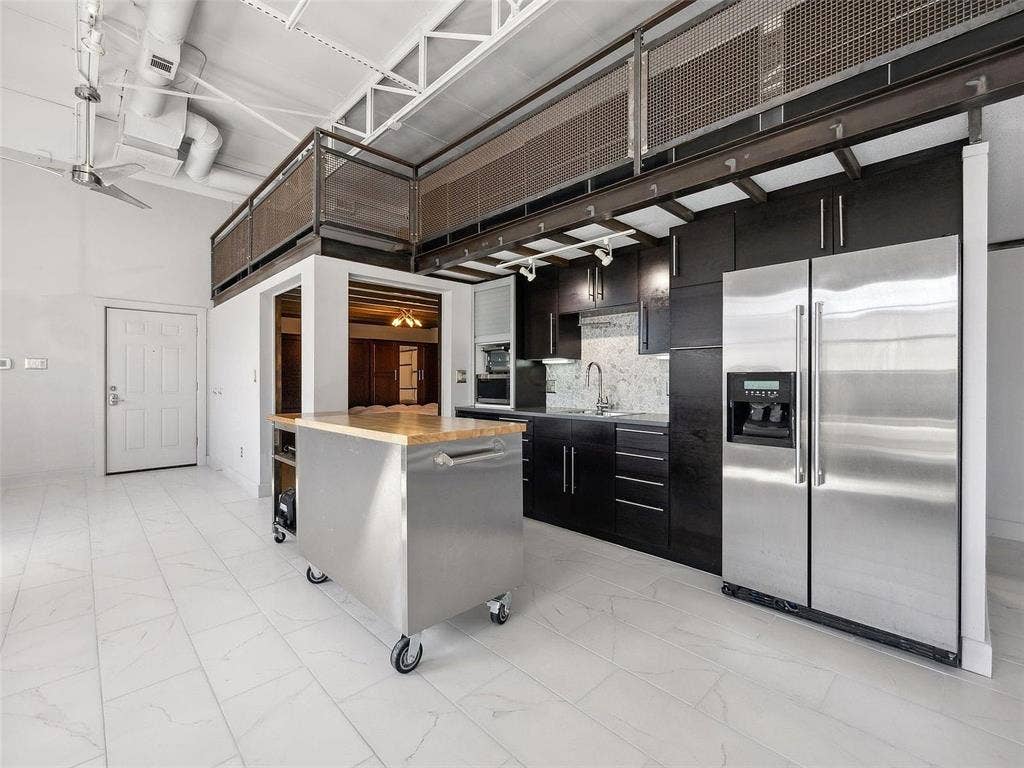 215 N Walton Street, #2, Dallas, TX 75226 Industrial-style loft featuring high ceilings with exposed trusses, dark wood cabinetry, a stainless steel kitchen island, tiled flooring, and a metal-mesh mezzanine railing