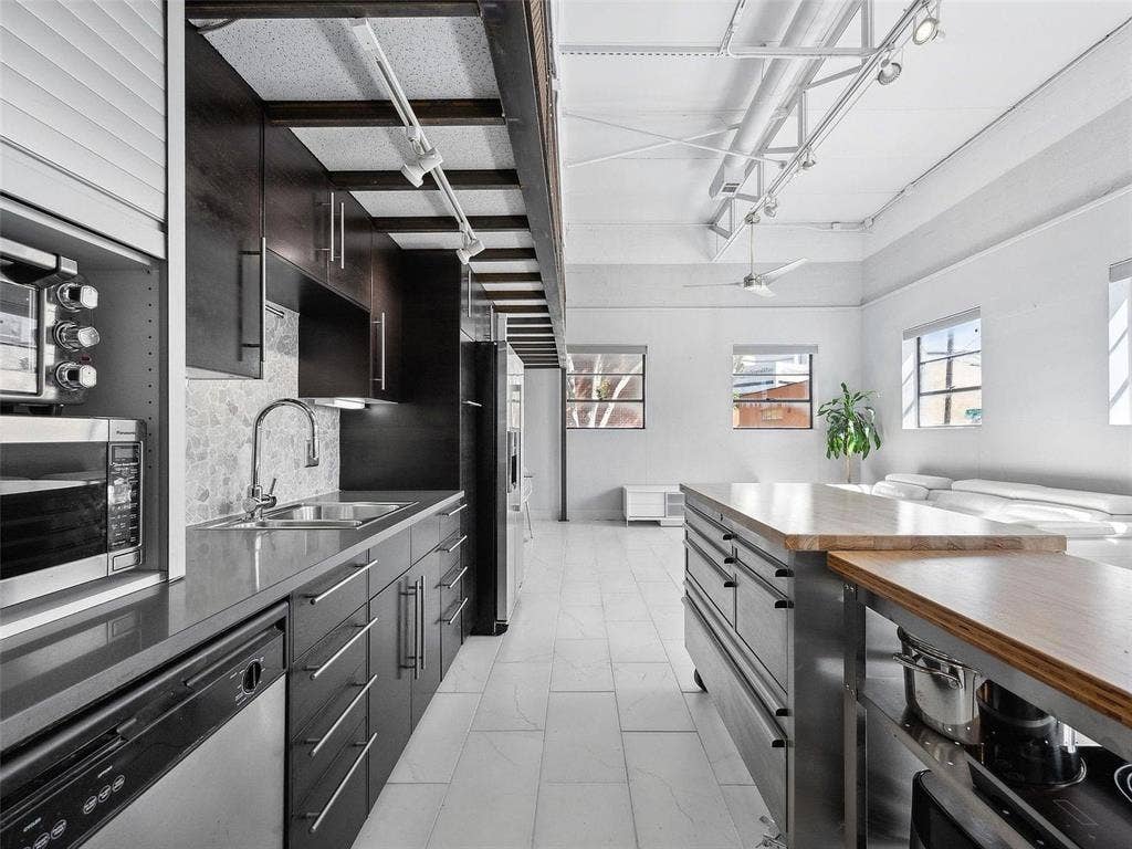215 N Walton Street, #2, Dallas, TX 75226 Industrial-style kitchen featuring sleek dark cabinetry, quartz countertops, tile flooring, exposed structural beams with track lighting, and a dual-level kitchen island with a wood-finish surface