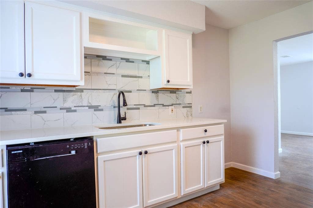 3229 Hardeman Street, Fort Worth, TX 76119 White shaker-style cabinetry, solid-surface countertop, matte black high-arch faucet, integrated dishwasher, and tiled backsplash featuring stone-patterned accents