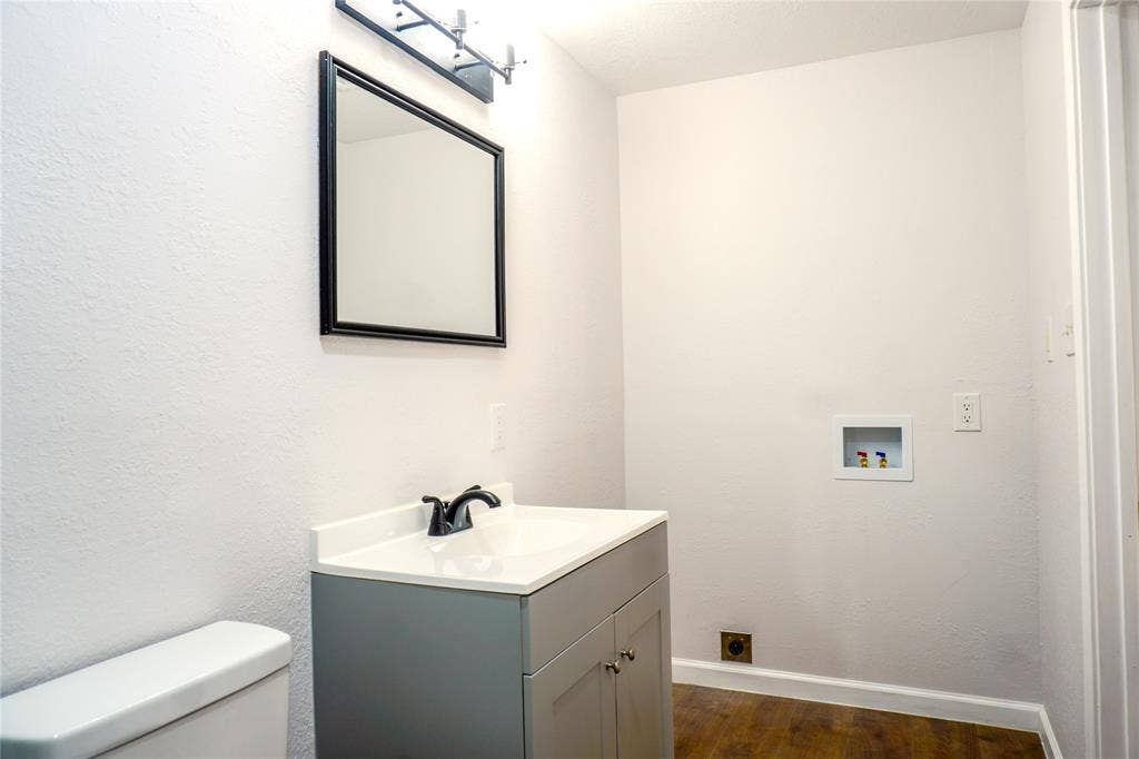3229 Hardeman Street, Fort Worth, TX 76119 Vanity featuring a gray cabinet with white countertop, integrated sink, and black faucet, complemented by a framed rectangular mirror, overhead lighting, and wood-finish flooring