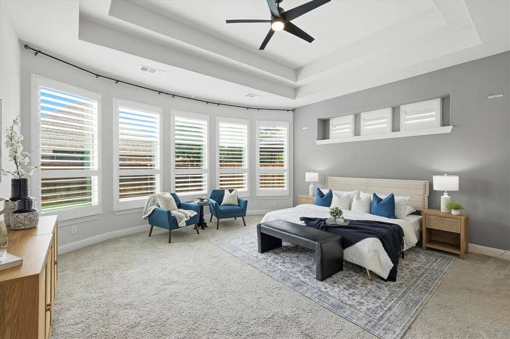 Spacious primary bedroom featuring a tray ceiling, expansive bay windows with plantation shutters, and reading area.