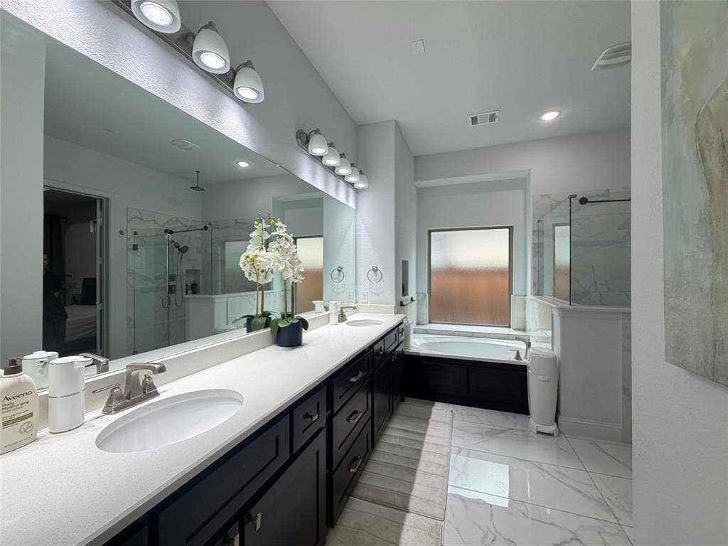 Dual-sink vanity with light-toned quartz countertops and dark cabinetry, large wall-mounted mirror, soaking tub, and walk-in glass-enclosed shower with tiled surround