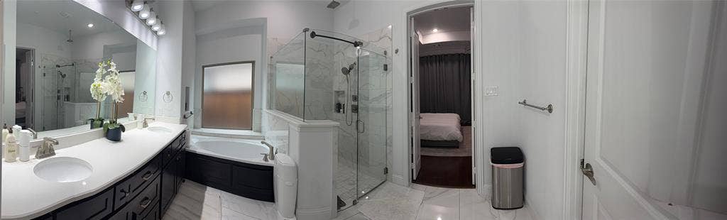 Double vanity with stone-style countertop and integrated sinks, deep soaking tub, glass-enclosed shower with built-in niche, and large wall-to-wall vanity mirror