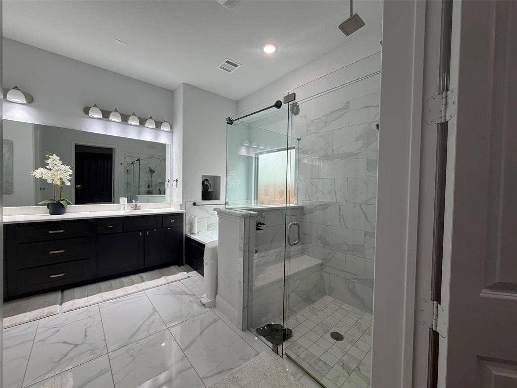 Bathroom featuring dark cabinetry with a light countertop, large-format tile flooring, a glass-enclosed shower with tiled walls and built-in bench, and a wall-to-wall mirror