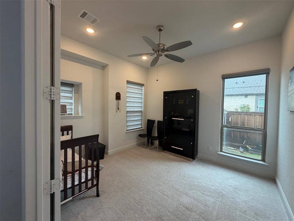 Neutral-toned carpeting, recessed ceiling lighting, a multi-blade ceiling fan, and multi-paned windows featuring dual-layer adjustable roller shades