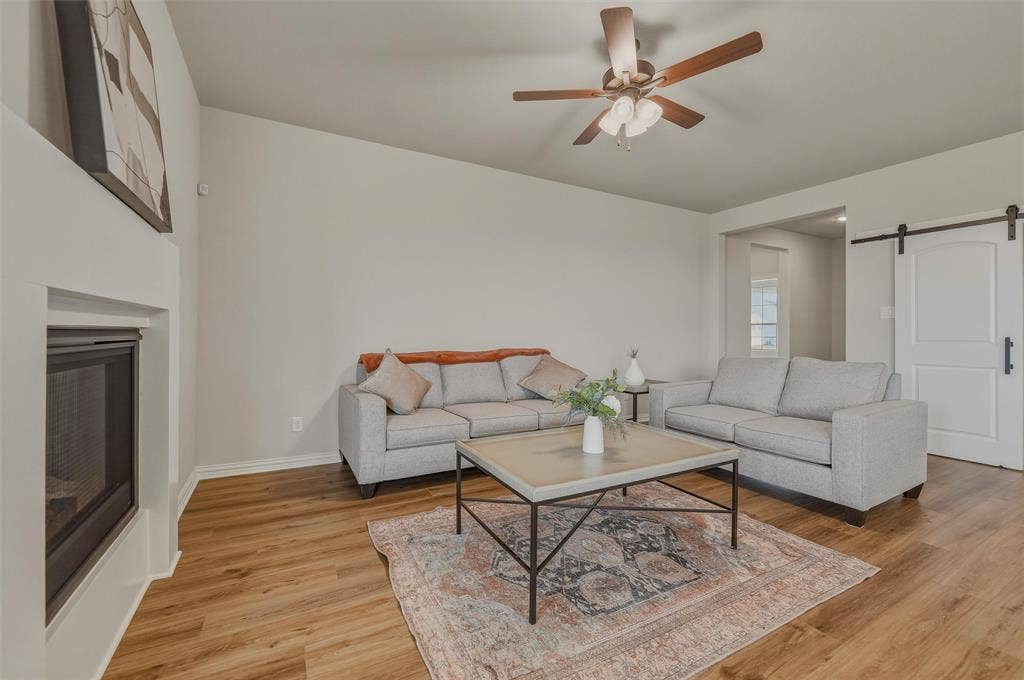Wood-finish flooring throughout, featuring a fireplace with a clean-lined mantel, a ceiling fan with wood-tone blades, and a barn door