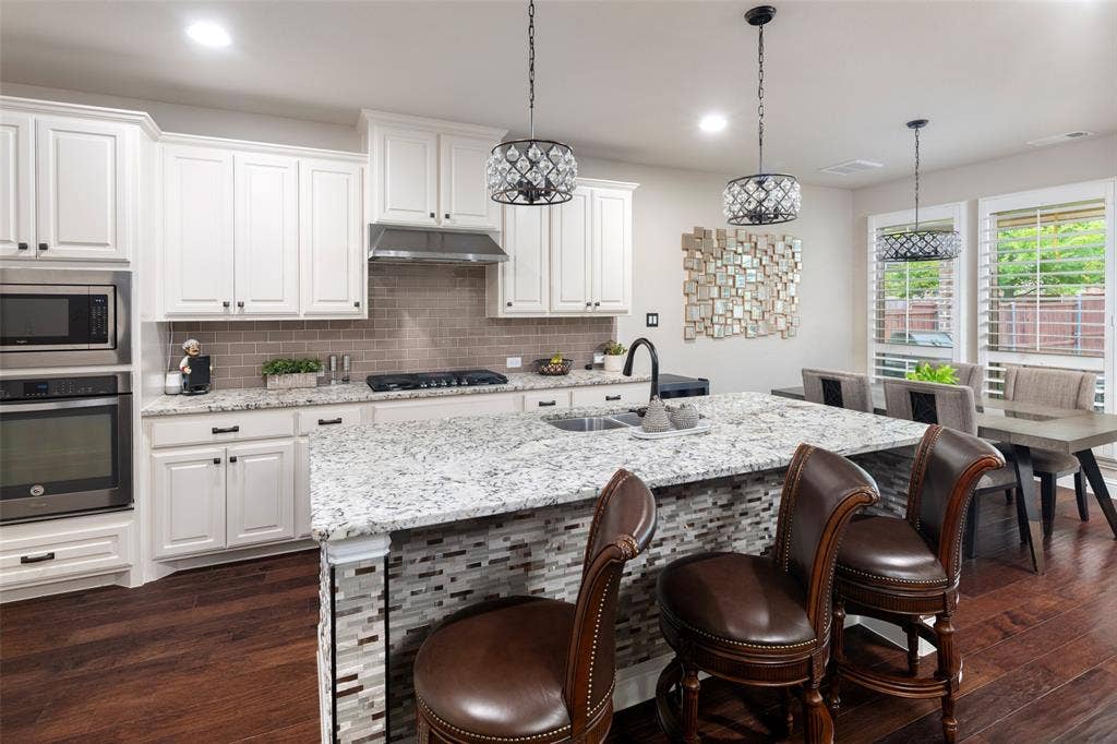 Kitchen featuring white cabinetry, stone countertops, stainless steel wall-mounted oven and microwave, dark wood-finish flooring, and a central island with decorative tile detailing