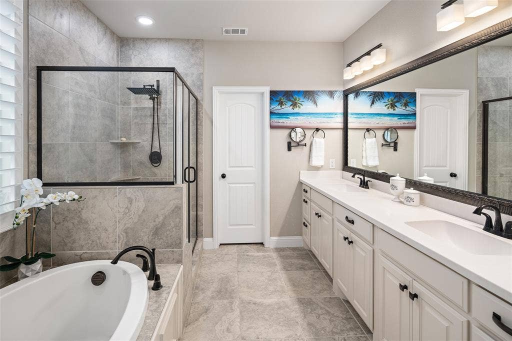 Dual-sink vanity with light-toned cabinetry and solid-surface countertop, freestanding soaking tub, enclosed glass-wall shower, and stone-finish tile flooring