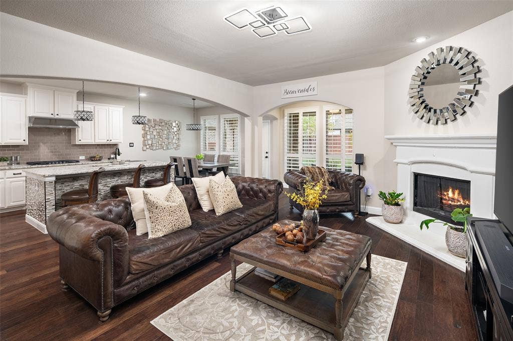 Open-concept living space featuring wood-finish flooring, a fireplace with a decorative mantel, white cabinetry, a kitchen island with a stone-finish countertop, and recessed ceiling lighting
