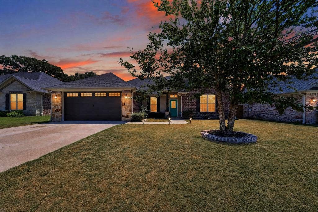 Single-story brick exterior featuring a multi-gabled roofline, an attached two-car garage, a paved driveway, and a stone-bordered tree planter