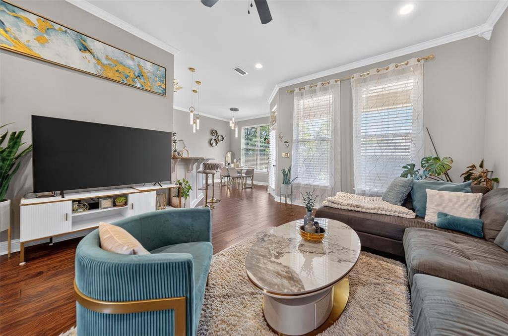 Open-concept interior featuring wood-finish flooring, crown molding, tiered pendant lighting, and multiple tall windows with decorative trim