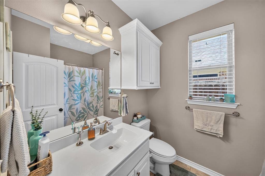 Bathroom featuring a white vanity cabinet with an integrated sink, brushed-finish faucet, wall-mounted upper cabinet, multi-bulb overhead light fixture, and a window with horizontal blinds