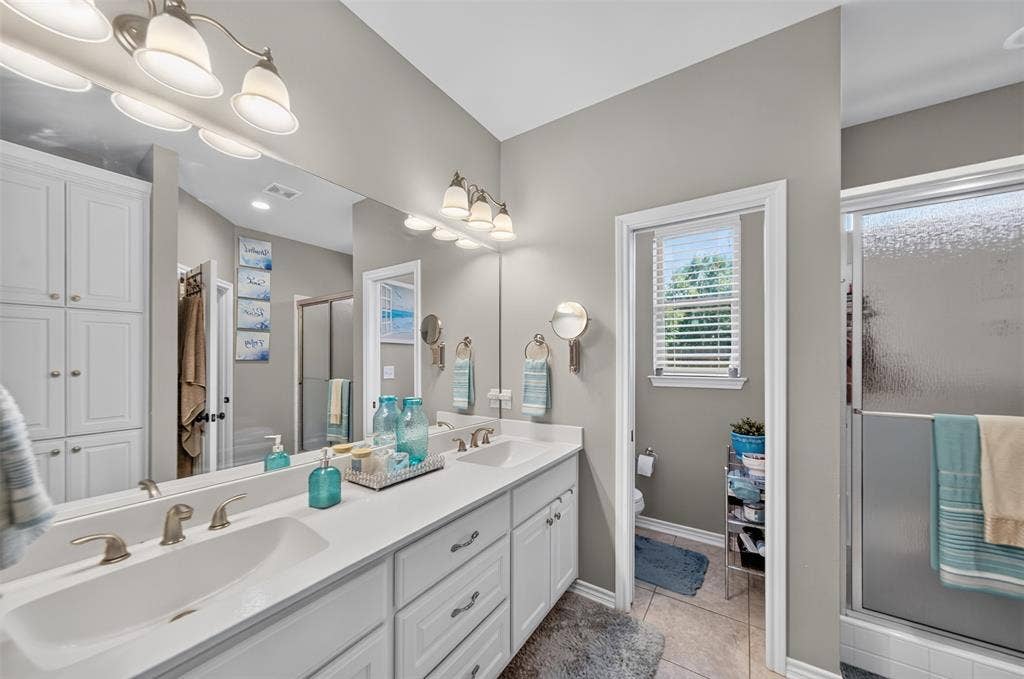 Dual-sink vanity with white cabinetry, light-toned solid surface countertops, polished metal fixtures, full-width wall mirror, and integrated storage cabinetry