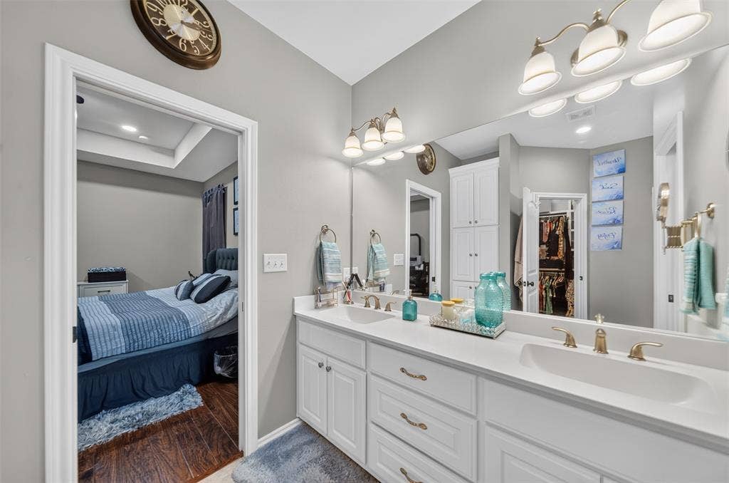 Double vanity featuring white cabinetry, integrated sinks, solid surface countertops, polished metal fixtures, and a wall-to-wall mirror with attached lighting