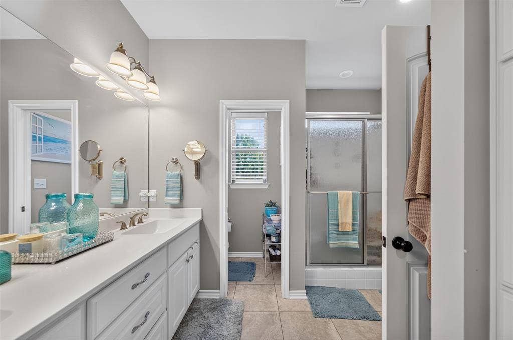 Bathroom featuring a white dual-sink vanity with stone countertop, four-light vanity fixture, expansive wall-mounted mirror, and a walk-in shower with glass sliding doors