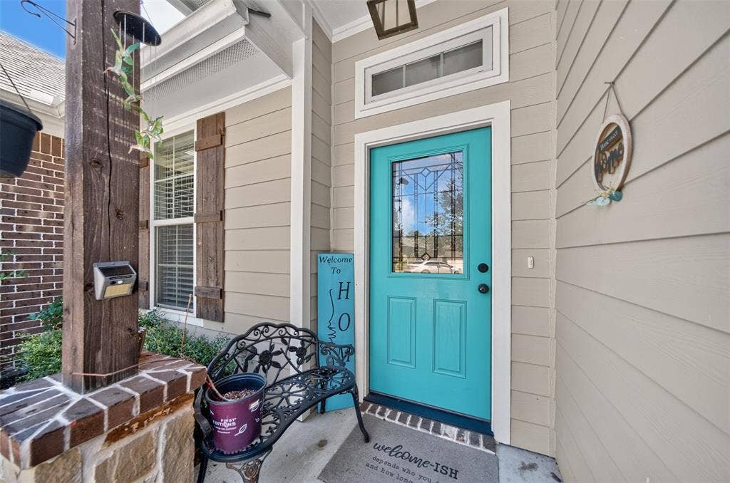 Covered entryway featuring a solid-panel door with a decorative glass insert, horizontal lap siding, a brick facade accent, wood-finish shutters, and a stone-base support column