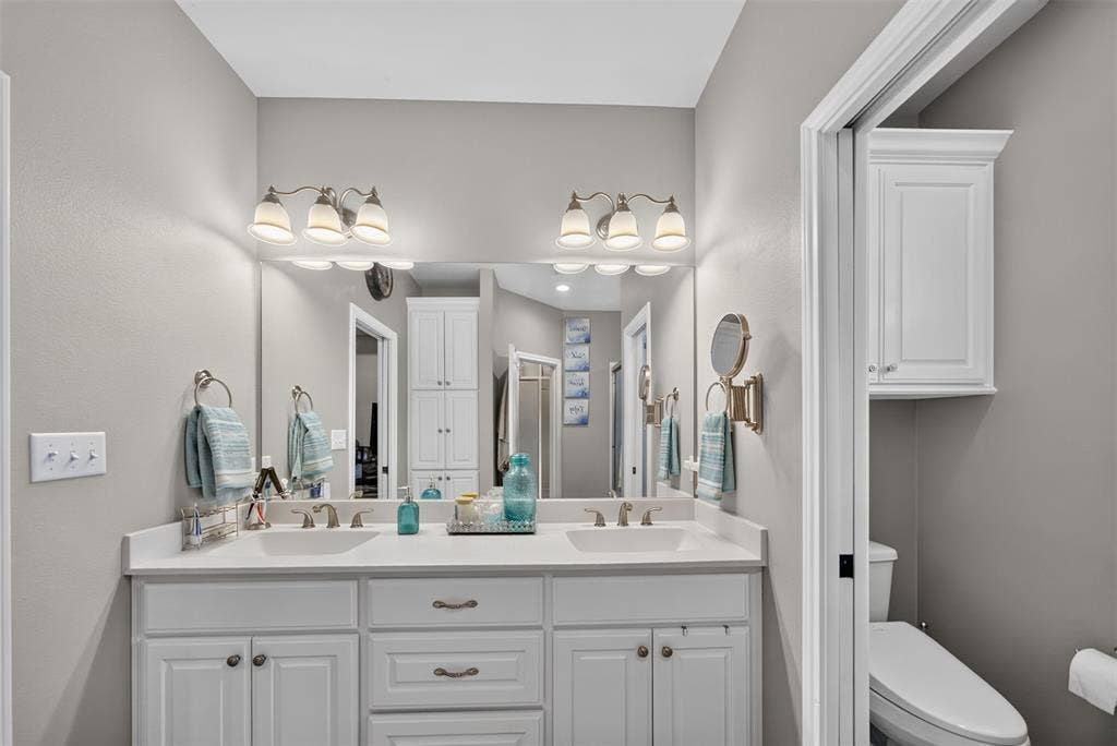Dual-sink vanity with white cabinetry, light-toned solid surface countertop, twin three-light wall fixtures, and integrated full-width mirror