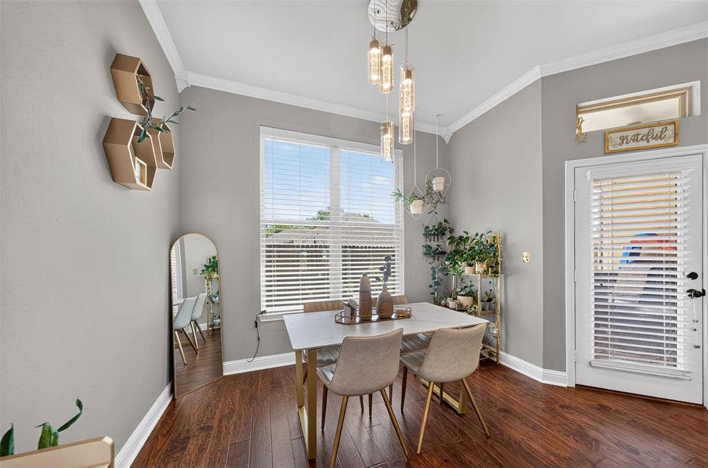 Room featuring wood-finish flooring, crown molding, multi-light pendant fixture, large window with plantation-style blinds, and exterior access door with transom window