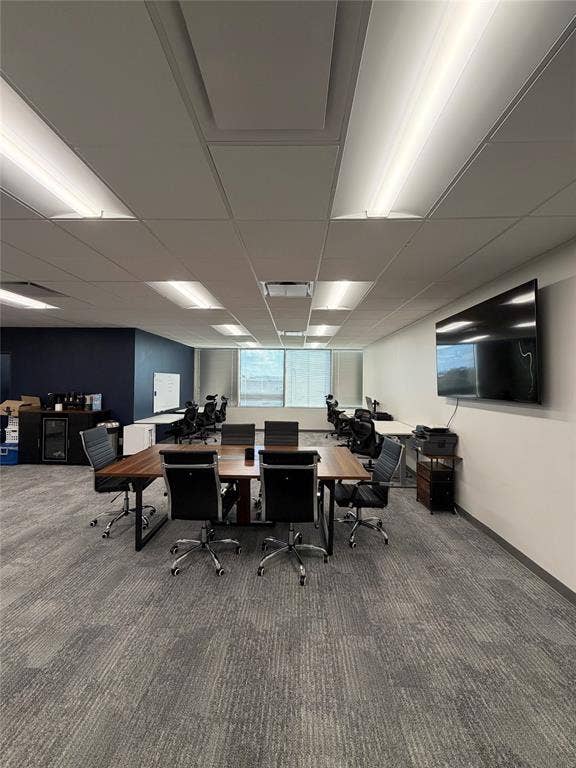 Interior space featuring a drop ceiling with recessed rectangular lighting, grey patterned carpeting, expansive window with natural light, and a built-in accent wall
