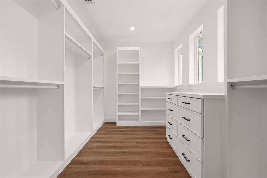 Walk-in storage space featuring white built-in cabinetry, custom shelving, integrated dresser drawers with matte black pulls, and wood-finish flooring