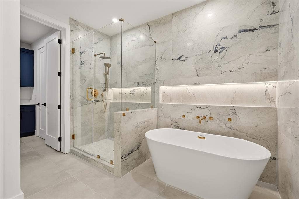 Freestanding oval soaking tub with metallic wall-mounted hardware, glass-enclosed walk-in shower with rainfall showerhead, floor-to-ceiling stone-patterned wall tiling, and integrated wall niches with accent lighting