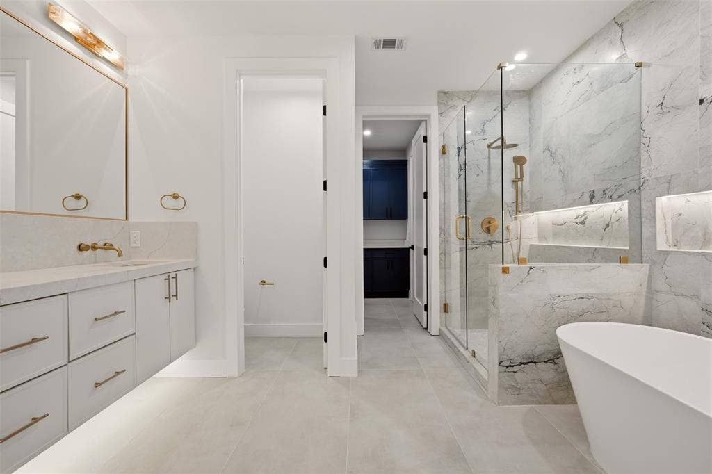 Floating vanity with stone countertop, freestanding soaking tub, glass-enclosed walk-in shower with stone surround, recessed shower niche, and brushed metallic fixtures