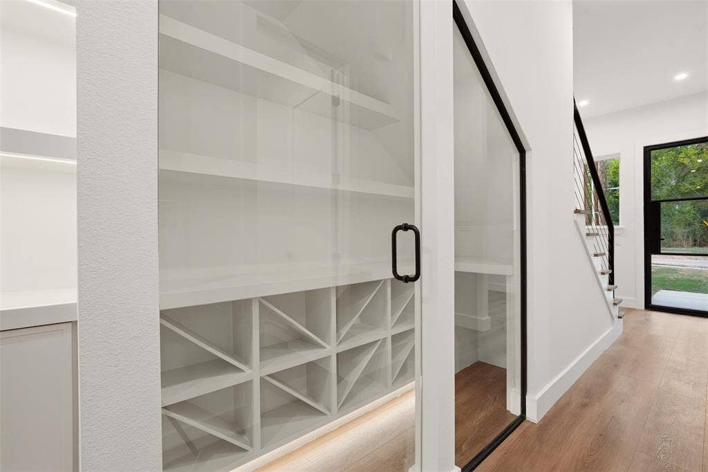 Built-in storage area featuring custom white shelving, integrated X-pattern wine rack, glass-paneled door with matte black hardware, recessed under-shelf lighting, and wood-finish flooring