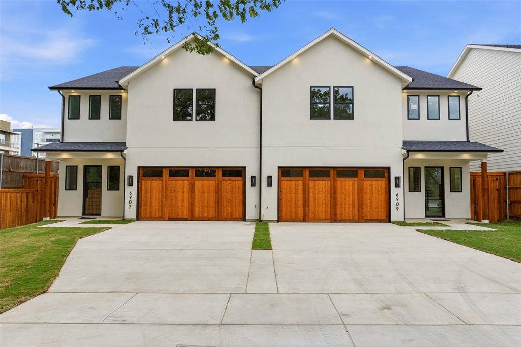 Duplex exterior featuring dual-pitched rooflines, smooth stucco siding, stained wood-finish garage doors with windows, and black-framed exterior lighting