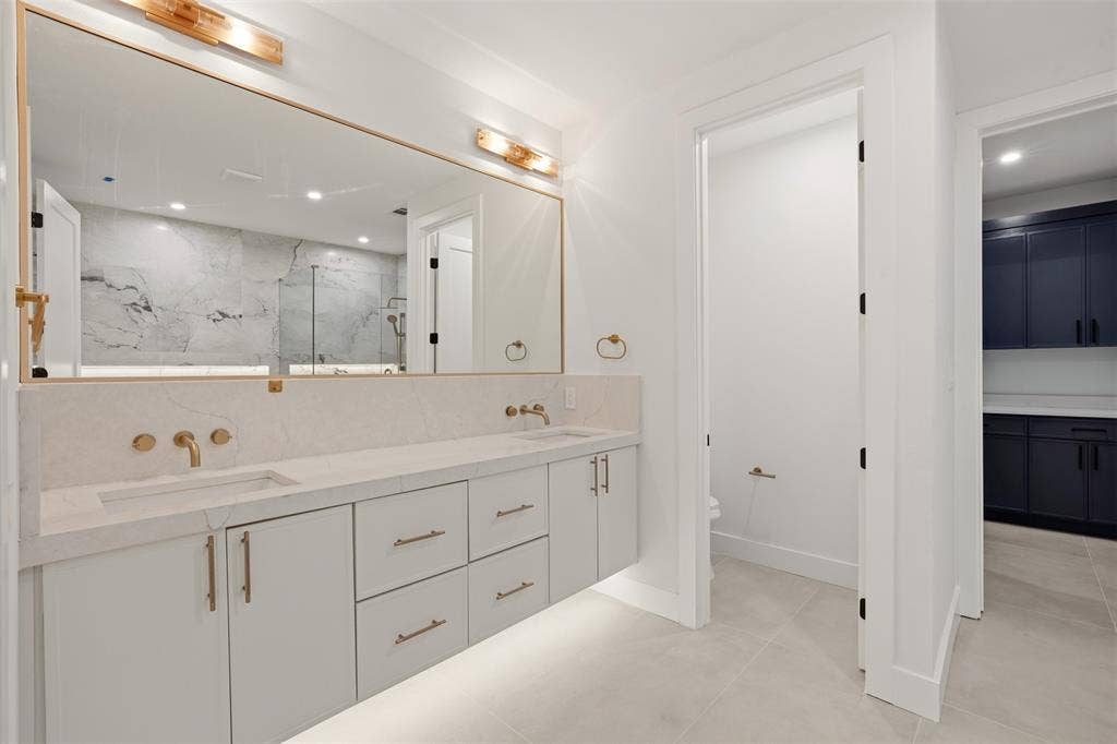 Floating vanity with stone countertop, dual undermount sinks, wall-mounted brass faucets, expansive framed mirror, and under-cabinet lighting