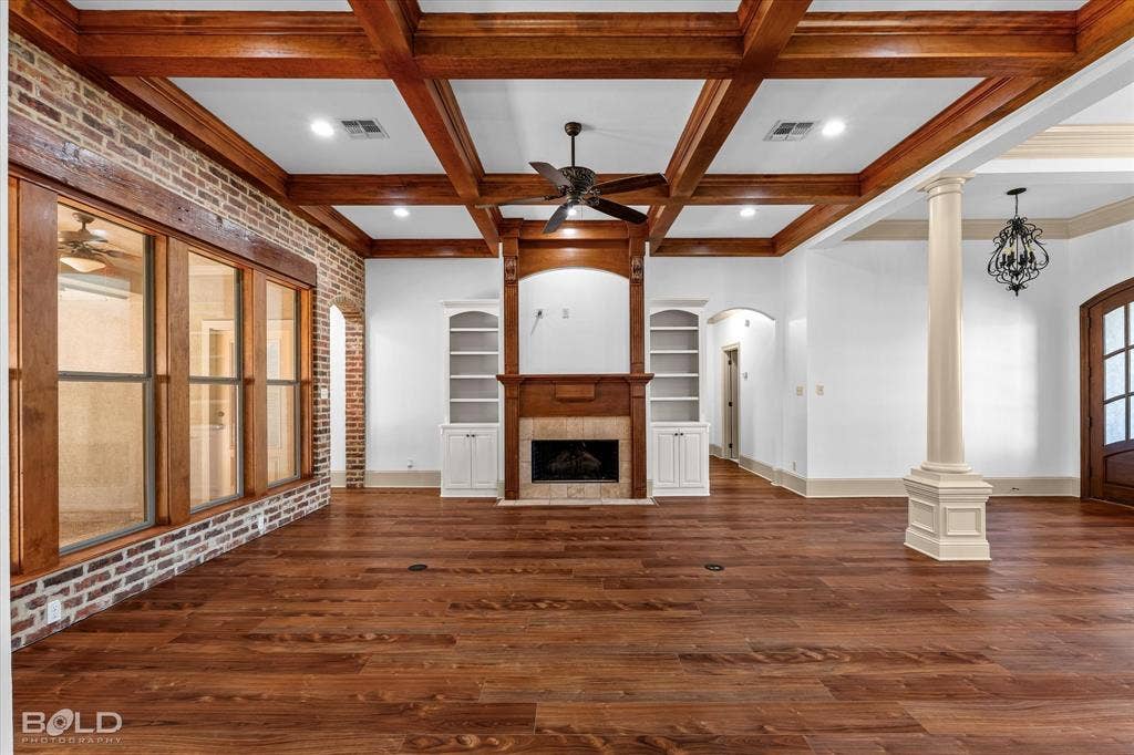 718 Duckwater Landing, Bossier City, LA 71111 Coffered wood-beam ceiling featuring a central ceiling fan, exposed brick wall, wood-finish flooring, built-in cabinetry flanking a fireplace with a stone-surround hearth, and a decorative fluted column