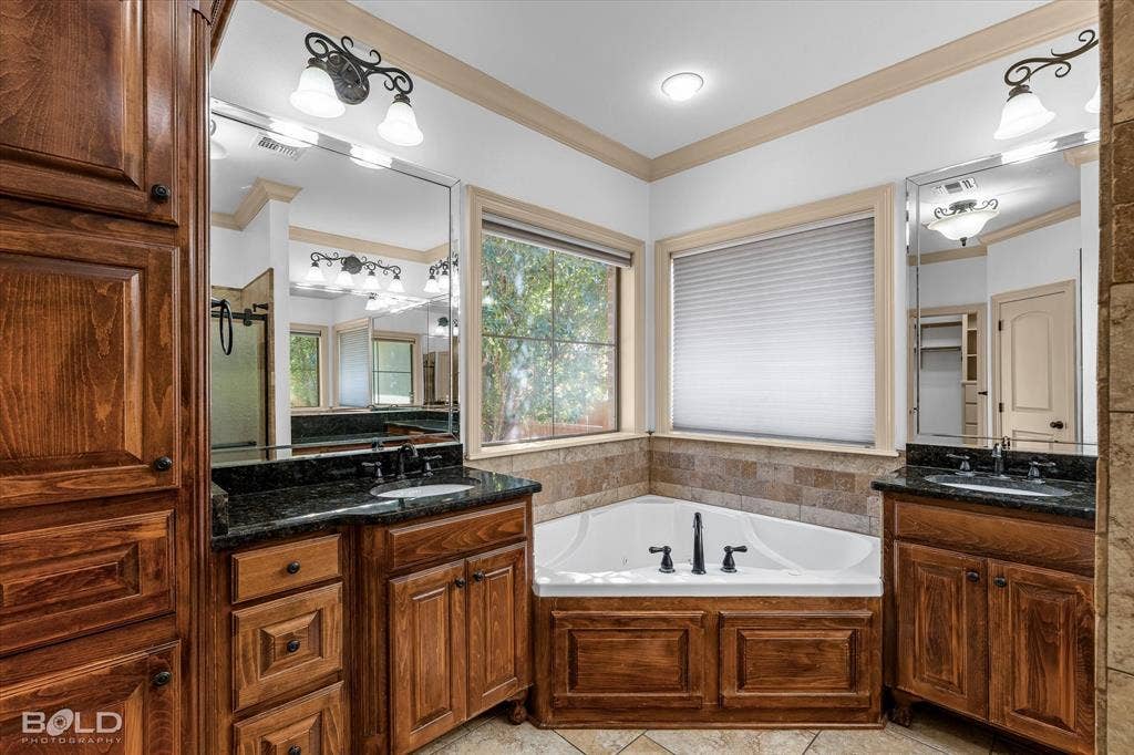 718 Duckwater Landing, Bossier City, LA 71111 Dual wood-finish vanities with stone countertops, corner soaking tub with tile surround, and wall-mounted light fixtures