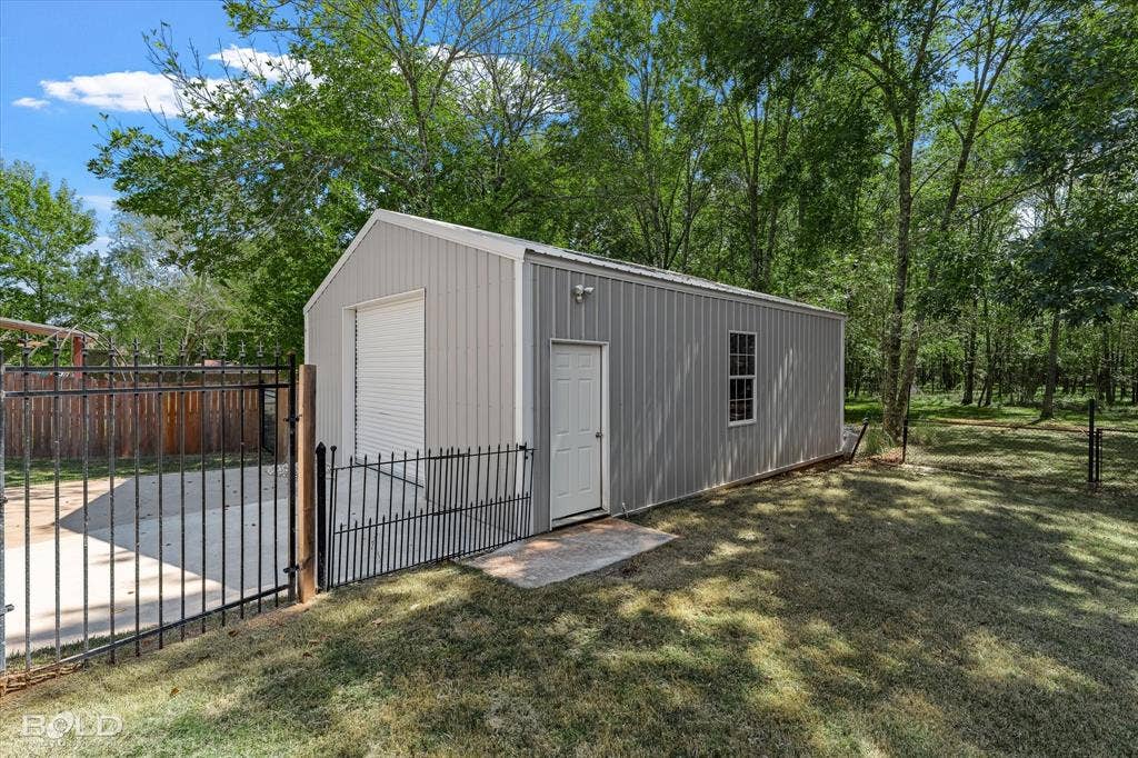 718 Duckwater Landing, Bossier City, LA 71111 Detached metal workshop featuring a roll-up bay door, pedestrian side entry door, single-pane window, and concrete apron, enclosed by a wrought-iron fence