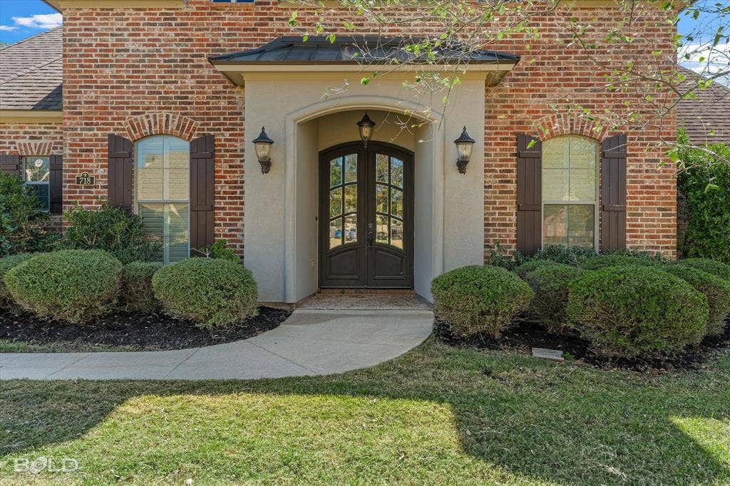 718 Duckwater Landing, Bossier City, LA 71111 Brick facade exterior featuring arched windows with decorative shutters, a covered entryway with a stucco surround, double glass-paneled front doors, and metal roof accenting