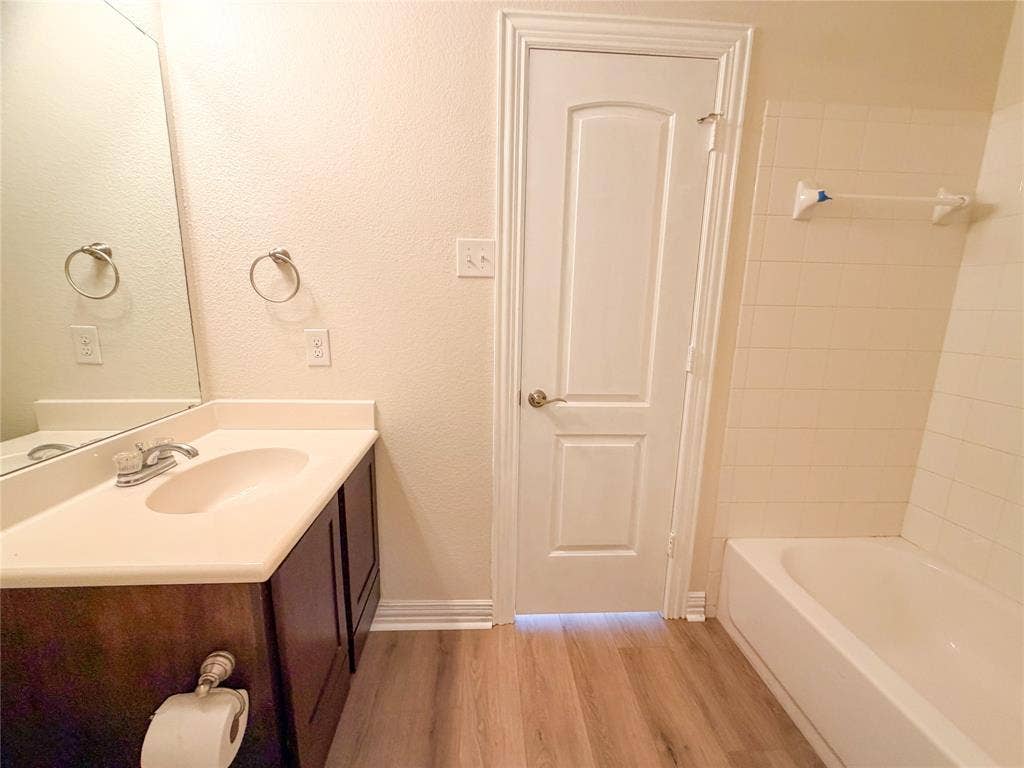 2410 S 2nd Street, #1373, Waco, TX 76706 Bathroom 1 featuring a single vanity with a white countertop, a built-in tub with white tile surround, a large wall mirror, and wood-finish flooring