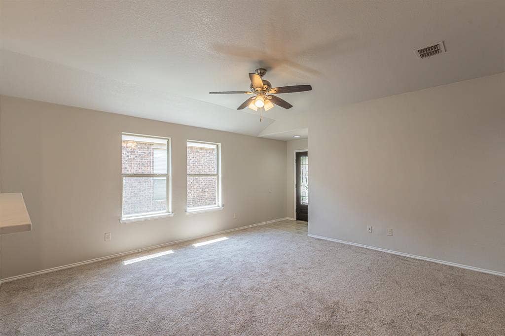 Carpeted room featuring light-colored walls, two double-hung windows, a ceiling fan with integrated lighting, and a dark-paneled door with decorative glass