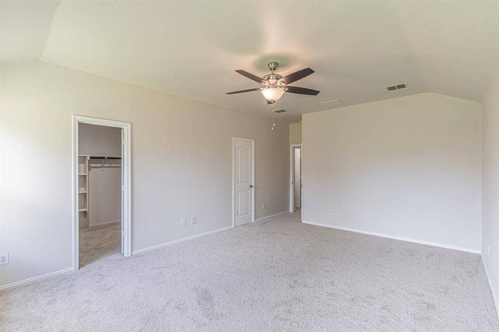 Spacious room featuring vaulted ceilings, light-toned carpeting, a ceiling fan with integrated lighting, and multiple doorways including one to a walk-in closet with built-in shelving