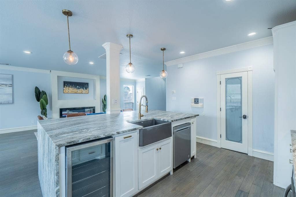 Kitchen island featuring waterfall-edge stone countertop, farmhouse-style sink, brass-finish faucet, built-in wine refrigerator, and pendant lighting