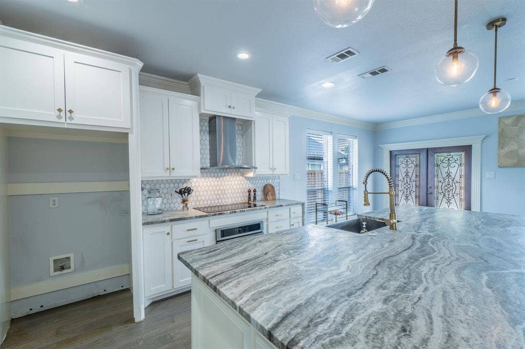 Kitchen featuring white shaker cabinetry, expansive stone-topped island with an integrated sink, geometric tile backsplash, stainless steel hood, and pendant lighting