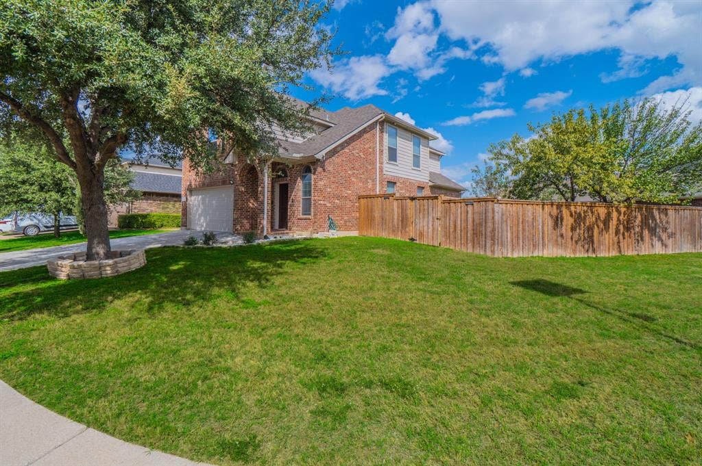 Two-story brick exterior featuring a multi-gable roofline, attached garage, stone-bordered tree planter, and wooden privacy fencing