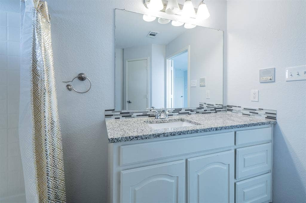 Bathroom vanity featuring white cabinetry, stone-finish countertop, integrated sink, tiled backsplash, and a wall-mounted light fixture
