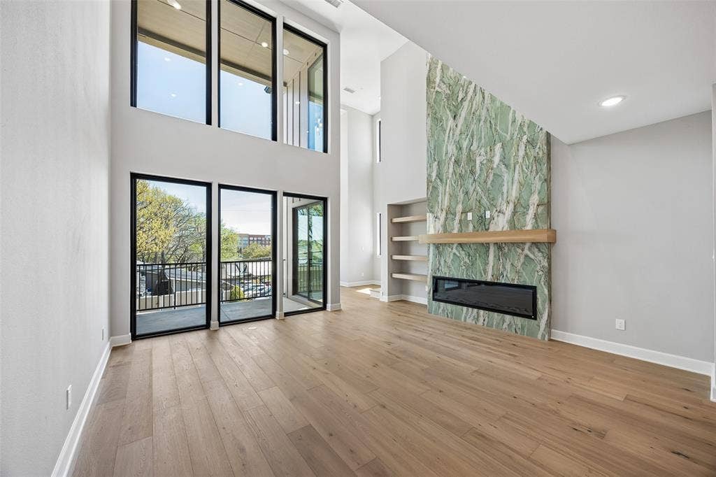 Double-height living area featuring floor-to-ceiling windows, wide-plank wood-finish flooring, a full-height stone fireplace surround with integrated hearth, and built-in wall shelving