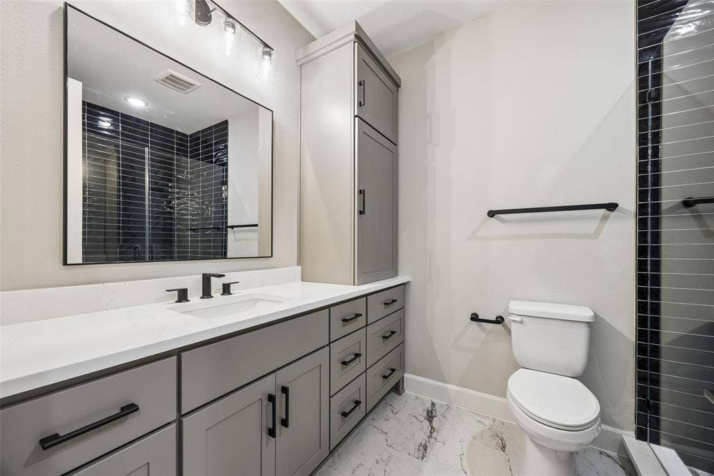 Grey shaker-style cabinetry with matte black hardware, white countertop with integrated sink, rectangular mirror with thin dark frame, multi-light vanity fixture, and patterned tile flooring
