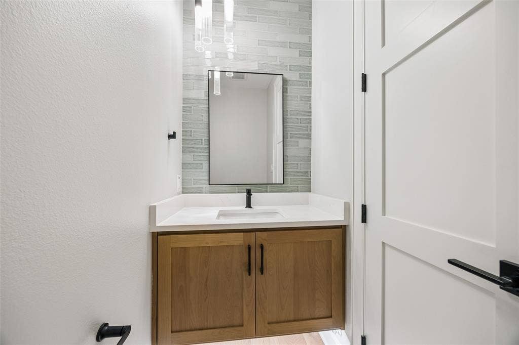Floating wood-finish vanity with a white countertop, integrated rectangular sink, matte black hardware, and linear tile backsplash accented by a framed mirror and pendant lighting