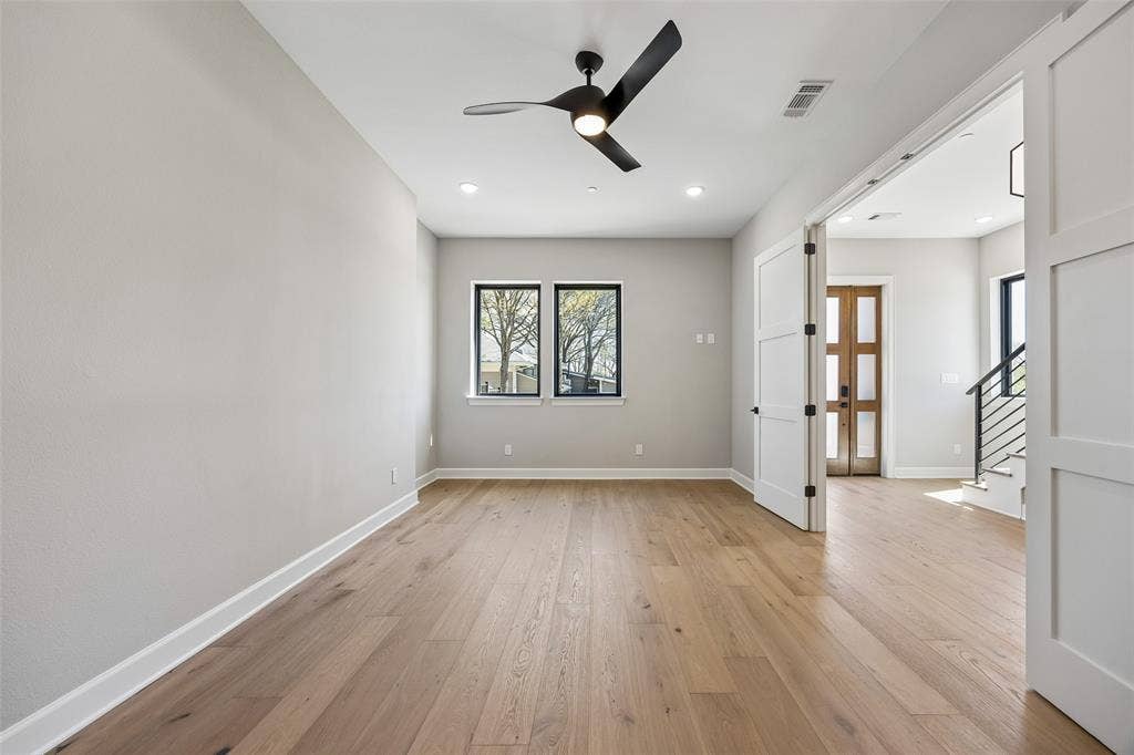 Light-filled interior space featuring wood-finish flooring, a modern black ceiling fan, dual windows with black frames, and white shaker-style double doors