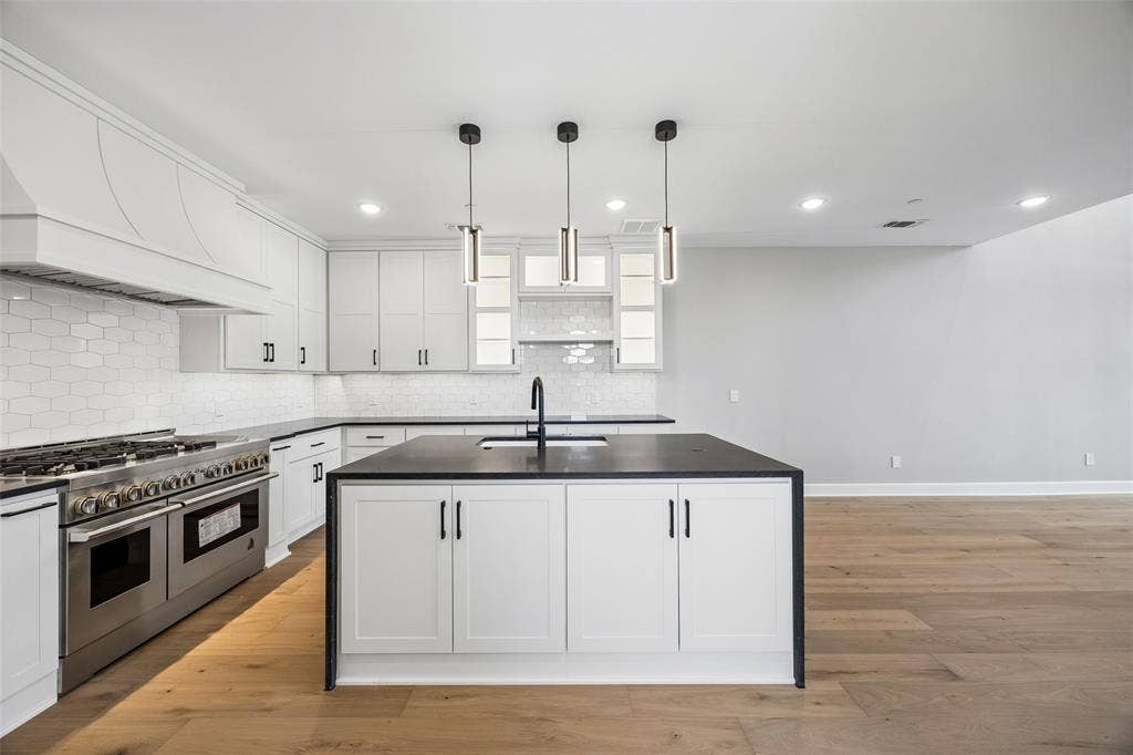 Kitchen featuring white cabinetry, a center island with a dark countertop and undermount sink, a stainless steel range with a custom hood, white hexagonal tile backsplash, and wood-finish flooring