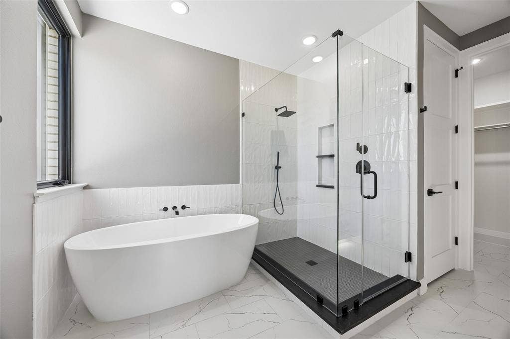 Freestanding soaking tub with wall-mounted faucet, walk-in glass-enclosed shower with matte black fixtures, built-in shower niche, and large-format tile flooring