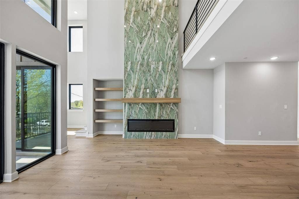 Double-height ceiling, floor-to-ceiling stone fireplace surround, linear built-in fireplace, wood-finish flooring, and minimalist floating shelving