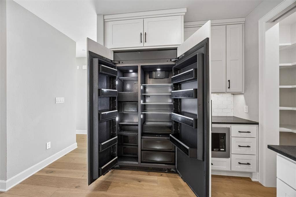 Built-in side-by-side refrigerator with interior lighting, shaker-style cabinetry, wood-finish flooring, dark solid-surface countertops, and tiled backsplash